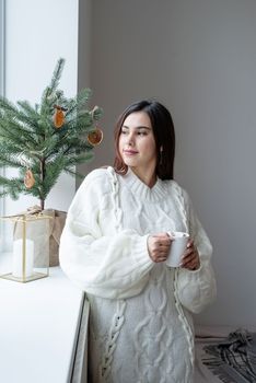 Merry Christmas and Happy New Year. Woman in warm white winter sweater standing next to the window at home at christmas eve holding cup with marshmallows, fir tree behind