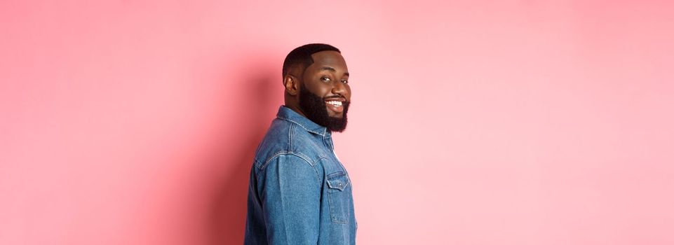 Handsome african-american man with beard, turn face at camera and smiling confident, standing over pink background.