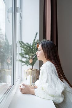 Merry Christmas and Happy New Year. Woman in warm white winter sweater lying in bed at home at christmas eve holding cup with marshmallows, fir tree behind