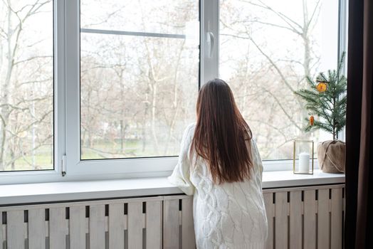 Merry Christmas and Happy New Year. Woman in warm white winter sweater lying in bed at home at christmas eve holding cup with marshmallows, fir tree behind