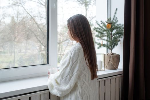 Merry Christmas and Happy New Year. Woman in warm white winter sweater standing next to the window at home at christmas eve holding cup with marshmallows, fir tree behind