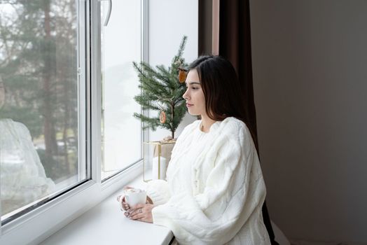 Merry Christmas and Happy New Year. Woman in warm white winter sweater lying in bed at home at christmas eve holding cup with marshmallows, fir tree behind