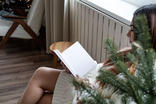Merry Christmas and Happy New Year. Woman in warm white winter sweater sitting next to the window at home at christmas eve reading book