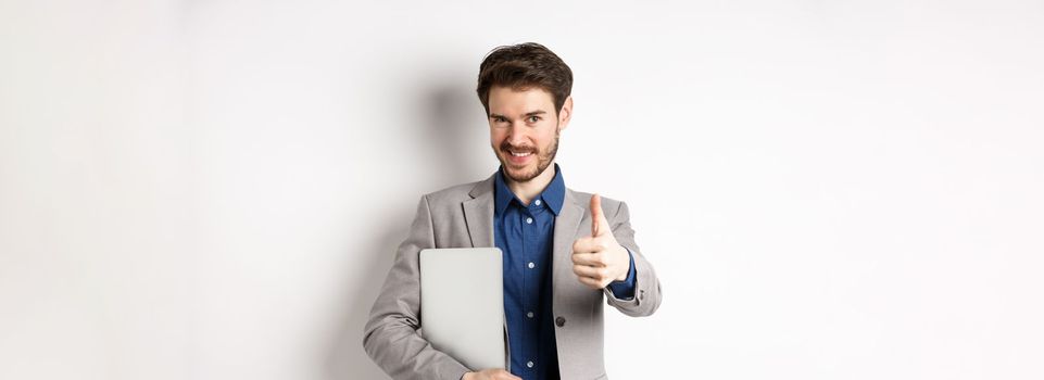 Successful businessman in stylish suit carry laptop and looking confident at camera, showing thumbs up in approval, white background.