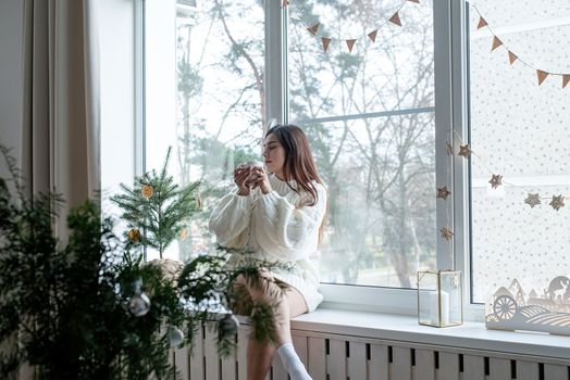 Merry Christmas and Happy New Year. Woman in warm white winter sweater lying in bed at home at christmas eve holding cup with marshmallows, fir tree behind