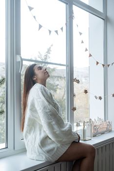 Merry Christmas and Happy New Year. Woman in warm white winter sweater sitting on the window at home at christmas eve holding cup with marshmallows, fir tree behind