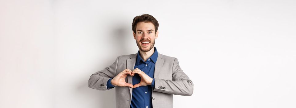 Passionate and romantic man in suit falling in love, showing heart sign and smiling at lover, standing on white background.