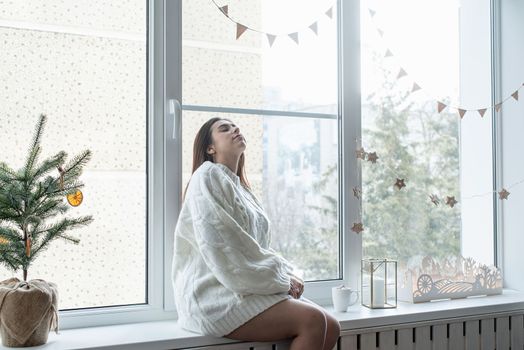 Merry Christmas and Happy New Year. Woman in warm white winter sweater sitting on the window at home at christmas eve holding cup with marshmallows, fir tree behind