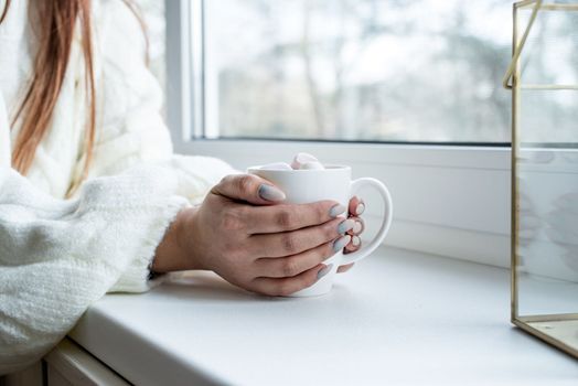 Merry Christmas and Happy New Year. Woman in warm white winter sweater sitting on the window at home at christmas eve holding cup with marshmallows, fir tree behind
