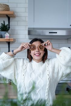 Merry Christmas and Happy New Year. Woman in warm white winter sweater standing at the kitchen at home with dry lemon slices next to face