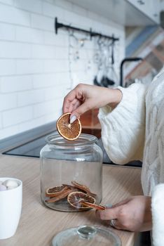 Merry Christmas and Happy New Year. Woman in warm white winter sweater standing at the kitchen at home making dry citrus decorations for christmas