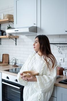 Merry Christmas and Happy New Year. Woman in warm white winter sweater standing at the kitchen at home holding bowl with popcorn