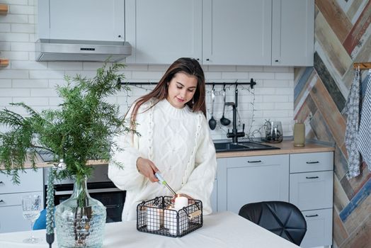 Merry Christmas and Happy New Year. Woman in warm white winter sweater standing at the kitchen at home lighting up the candles