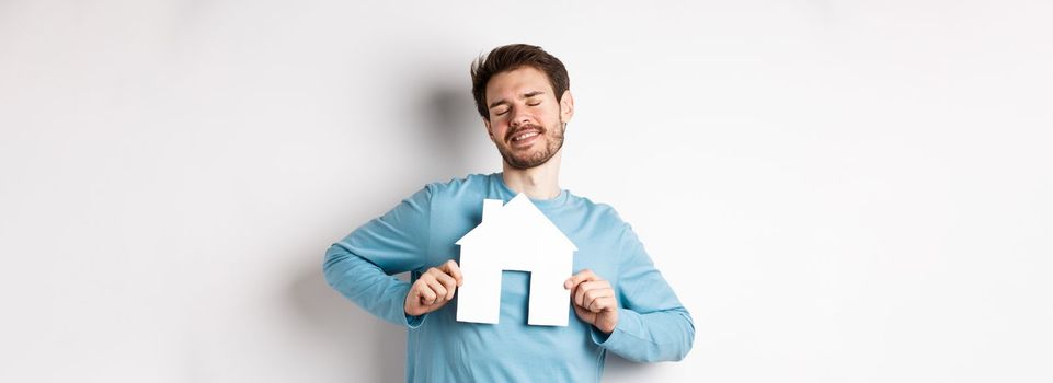 Real estate and insurance concept. Dreamy young man smiling with closed eyes, showing paper house cutout, wishing to buy home, standing over white background.