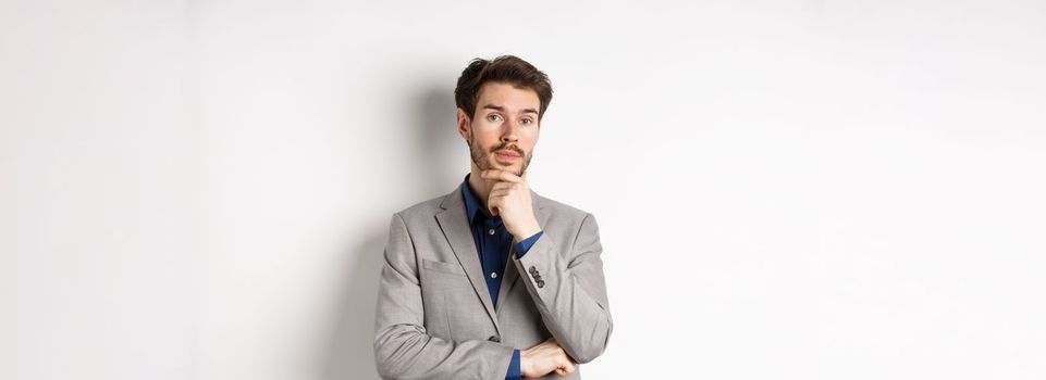 Thoughtful businessman in suit touching beard, thinking and looking at camera, deciding with pensive face, standing on white background.