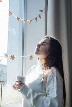 Merry Christmas and Happy New Year. Woman in warm white winter sweater sitting on the window at home at christmas eve holding cup with marshmallows, fir tree behind