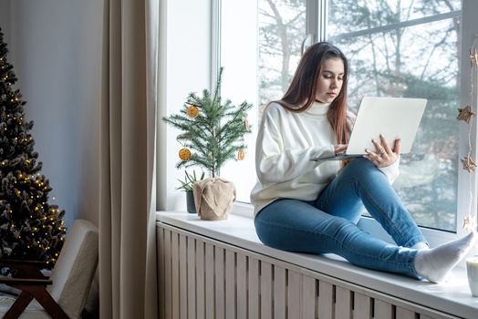 Merry Christmas and Happy New Year.Woman in warm white winter sweater sitting at windowsill at home at christmas eve working on laptop, fir tree behind