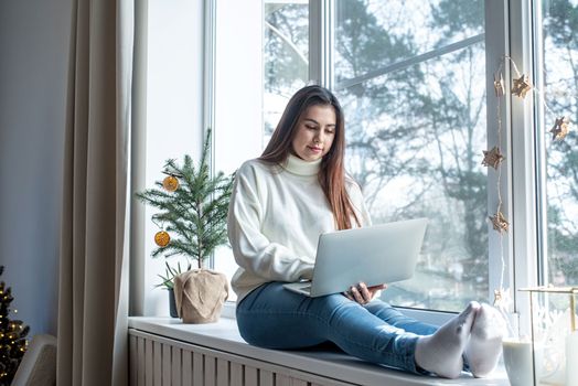 Merry Christmas and Happy New Year.Woman in warm white winter sweater sitting at windowsill at home at christmas eve working on laptop, fir tree behind