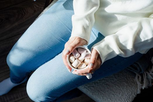 Merry Christmas and Happy New Year. Woman in warm white winter sweater holding cup with marshmallows, fir tree behind