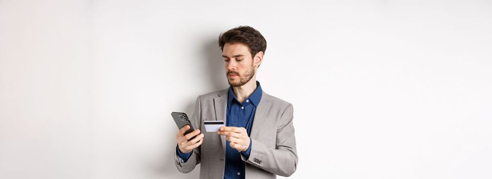 Online shopping. Serious man paying with credit card on smartphone, sending money, standing in suit on white background.