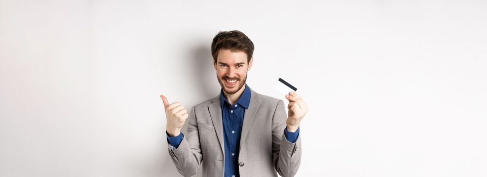 Happy bank client in business suit showing plastic credit card and thumb up, smiling satisfied, standing on white background.