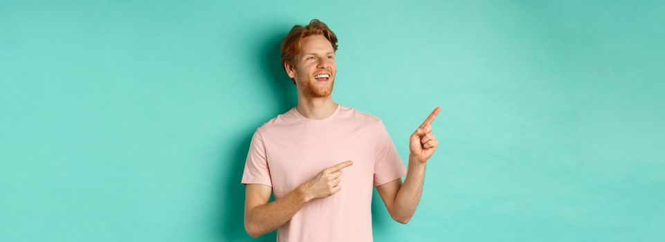 Dreamy handsome guy with red hair, smiling and looking at upper left corner with awe, admire something, standing over mint background. Copy space