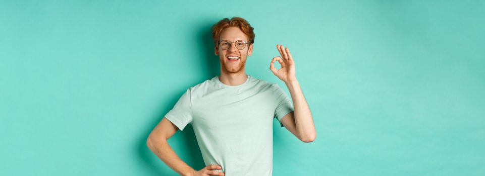 Happy young man with red hair and beard, wearing glasses and t-shirt, smiling satisfied and showing ok sign, say yes, approve and agree, standing over turquoise background.