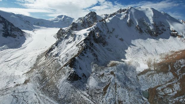 An ancient snow glacier among high mountains. The peaks of the mountains are covered with snow. In places there are steep cliffs and large rocks. Blue sky with white clouds. Top view from a drone