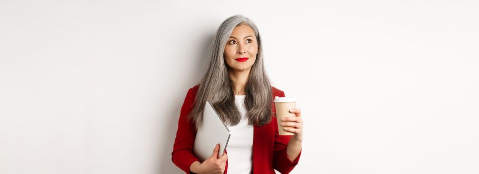 Business. Successful asian businesswoman with grey hair, drinking coffee and standing with laptop, looking thoughtful upper left corner.
