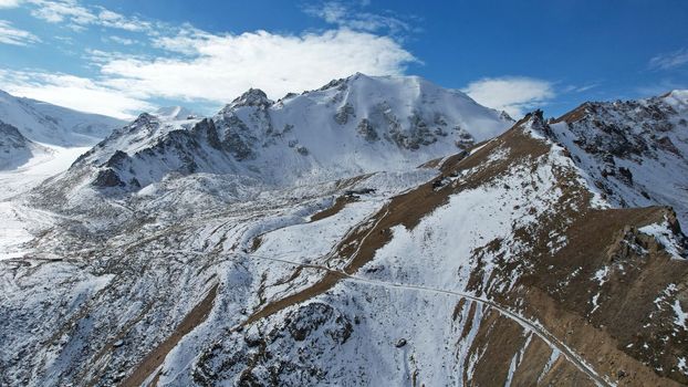 High snowy peaks and a small glaciological station. Drone view of the blue sky with clouds, steep cliffs and rocks. An ancient glacier covered with snow. Small houses are standing. Moraine lake