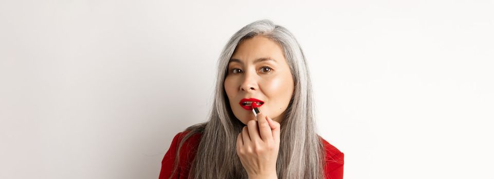 Beauty and makeup concept. Stylish asian mature woman with grey hair, looking in mirror and apply red lipstick, standing over white background.