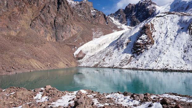 A mountain lake with emerald water reflects a glacier like a mirror. You can see the peaks of the mountains. The lake is partially frozen. There are large stones and snow in places. Moraine Lake