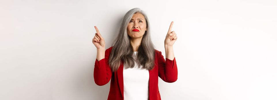 Disappointed asian senior woman looking, pointing fingers up and grimacing, dislike something, standing skeptical over white background.