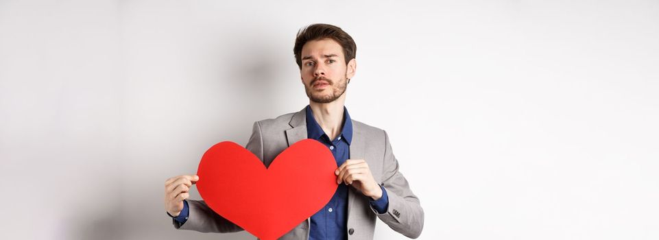 Passionate handsome man showing heart pounding gesture with red valentines cutout, standing in suit and searching for love, standing over white background.