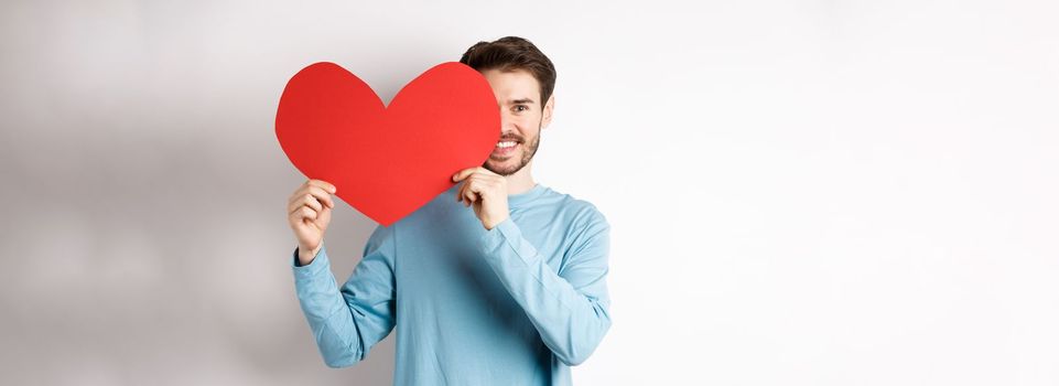 Smiling handsome man holding romantic red heart over half of face, surprise girlfriend on Valentines day, making love confession, standing over white background.