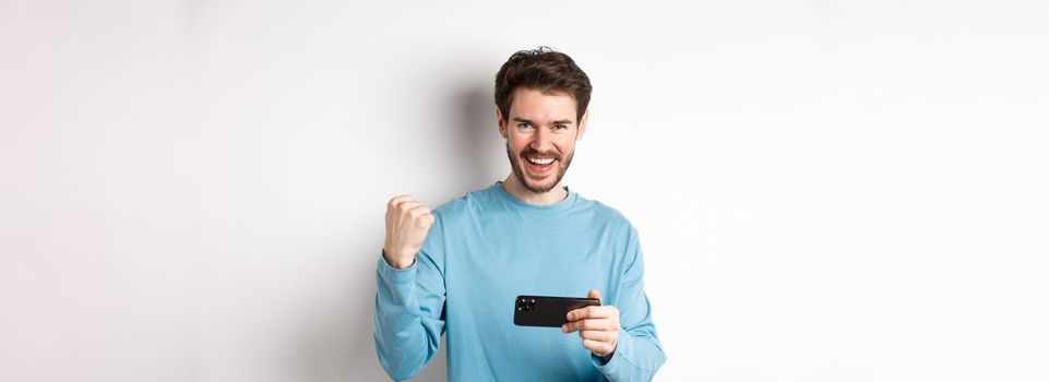 Lucky man winning in mobile video game, holding smartphone and saying yes, triumphing with fist pump, standing over white background.