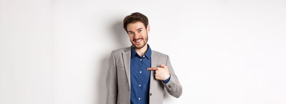 Modest smiling man in suit pointing at himself with cute face, self-promoting, standing on white background.