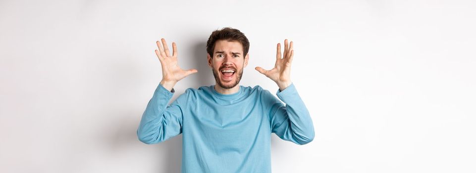 Image of freaked out young man screaming and shaking hands, shouting at camera frustrated or worried, panicking over white background.