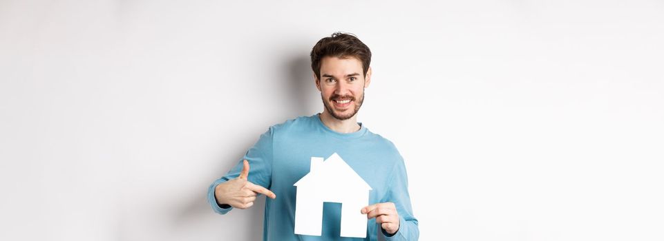 Real estate and insurance concept. Smiling young man pointing at paper house cutout and looking happy, buy apartment, standing over white background.