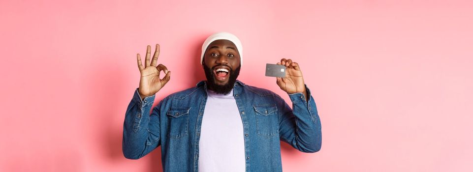 Shopping concept. Handsome african-american man recommending bank, showing credit card and okay sign, smiling satisfied, standing over pink background.