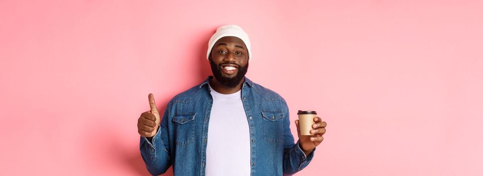 Handsome african-american hipster man showing thumb up, drinking coffee and recommending cafe, standing over pink background.