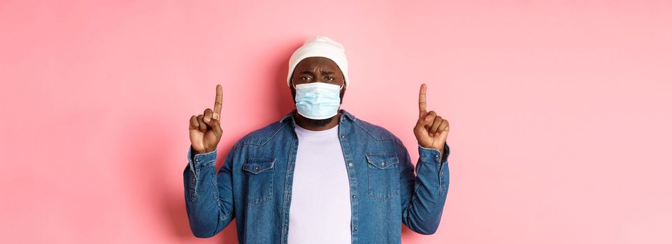 Coronavirus, lifestyle and global pandemic concept. Upset Black man in face mask frowning, pointing fingers up, showing advertisement, standing over pink background.