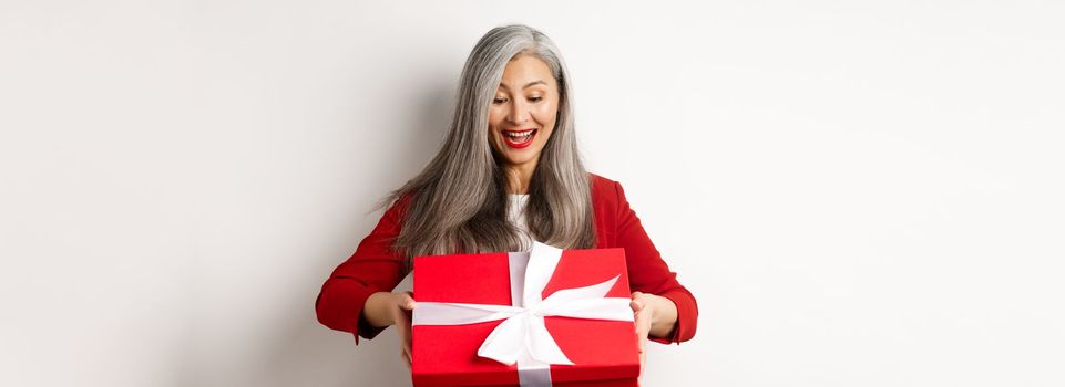 Happy elderly woman with grey hair, receive present, looking at red gift box and smiling surprised, standing over white background.