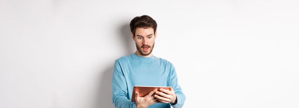 Image of man looking curious at digital tablet, reading interesting news online, standing on white background. Copy space