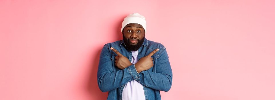 Confused Black man asking help with choice, pointing sideways and staring at camera, making decision, standing over pink background.