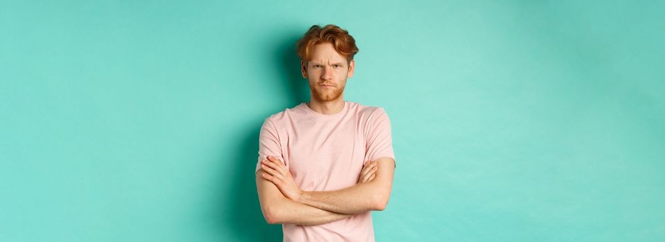 Gloomy redhead guy feeling offended, frowning upser with arms crossed on chest, looking insulted and sulking, standing over mint background.