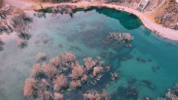 Turquoise blue mirror water with trees in the lake. Light streaks from underwater streams are visible. Autumn mountains and coniferous trees are reflected in the water. Issyk Mountain Lake, Kazakhstan