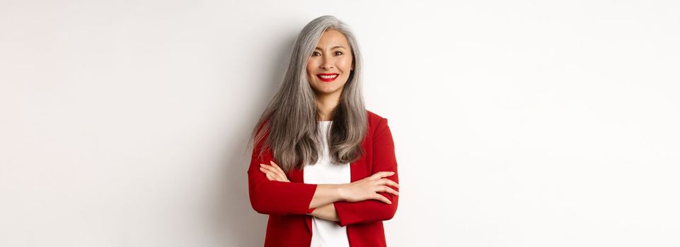 Business people. Smiling asian senior woman in red blazer, cross arms on chest and looking professional, standing over white background.