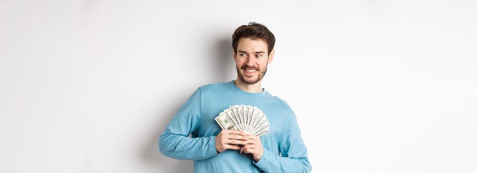 Greedy smiling man showing money and looking right, thinking about shopping, standing over white background.