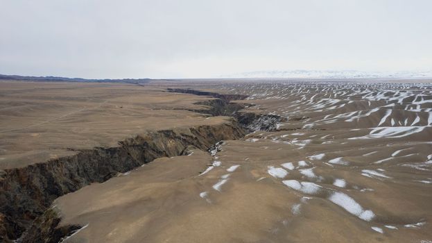A large crack in the ground overlooking the snowy hills. Grey sky covered with clouds. The Grand Canyon in the middle of the steppe. The river runs. Top view from a drone. Kazakhstan, Black Canyon
